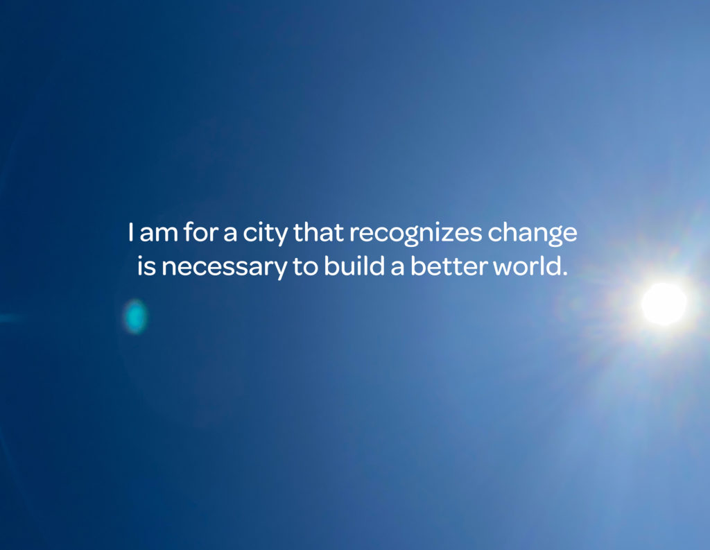 On a background of a clear blue sky, white text reads, "I am for a city that recognizes change is necessary to build a better world."